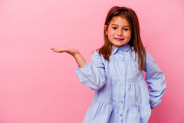Pequena menina caucasiana isolado no fundo rosa mostrando um espaço de cópia em uma palma e segurando outra mão na cintura. - Foto, Imagem