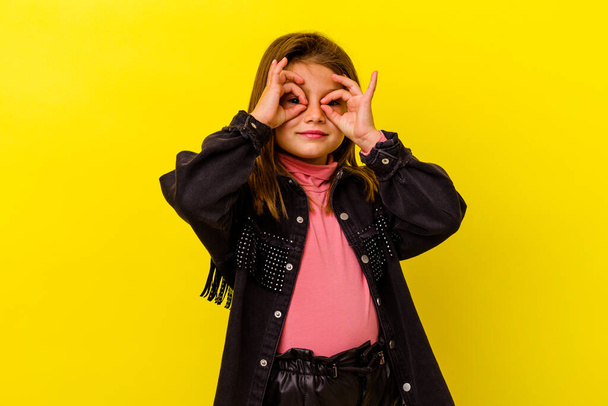 Little caucasian girl isolated on yellow background showing okay sign over eyes - Фото, изображение