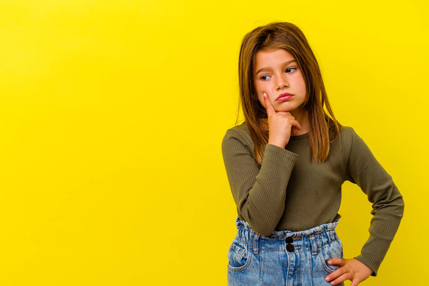 Pequena menina caucasiana isolado no fundo amarelo olhando para os lados com expressão duvidosa e cética. - Foto, Imagem