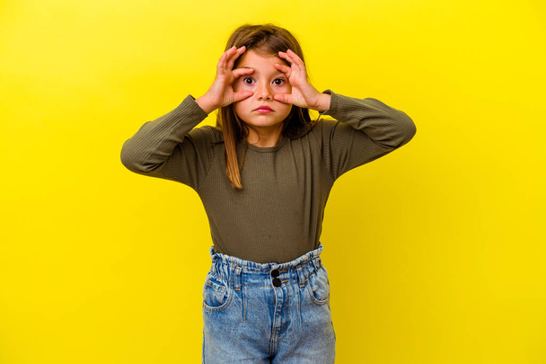 Petite fille caucasienne isolée sur fond jaune gardant les yeux ouverts pour trouver une opportunité de succès. - Photo, image