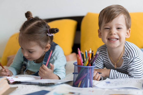 έξυπνο χαμογελαστό preshool μικρά παιδιά αγόρι και κορίτσι ζωγραφίζουν με μολύβια ξαπλωμένα στο στομάχι τους στο κίτρινο κρεβάτι. Μικρή χαριτωμένο αναγνώστη έχουν τη διασκέδαση, χαρούμενος παιδί σε καραντίνα στο σπίτι. Φιλία, οικογένεια, εκπαίδευση - Φωτογραφία, εικόνα