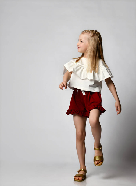 cute smiling little girl in a white blouse with flounces, burgundy shorts and sandals, walks briskly and looks to the side. - Fotó, kép