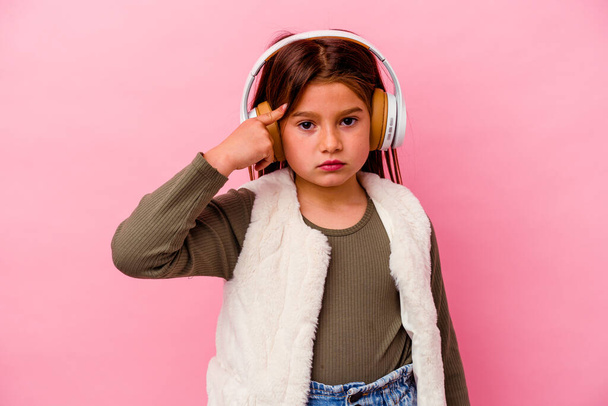 Little caucasian girl listening music isolated on pink background pointing temple with finger, thinking, focused on a task. - Foto, Imagen