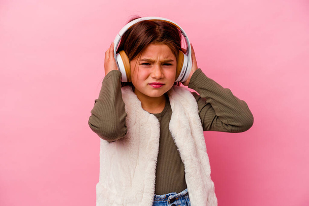 Little caucasian girl listening music isolated on pink background covering ears with hands. - Zdjęcie, obraz