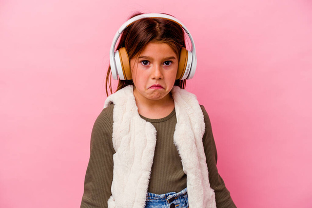Little caucasian girl listening music isolated on pink background shrugs shoulders and open eyes confused. - Foto, imagen