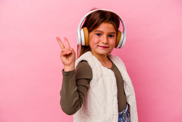 Petite fille caucasienne écoutant de la musique isolée sur fond rose joyeuse et insouciante montrant un symbole de paix avec les doigts. - Photo, image