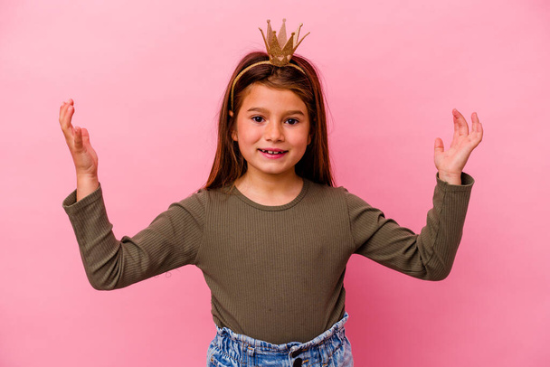 Μικρή πριγκίπισσα κορίτσι με στέμμα απομονωμένο σε ροζ φόντο λαμβάνει μια ευχάριστη έκπληξη, ενθουσιασμένος και σηκώνοντας τα χέρια. - Φωτογραφία, εικόνα