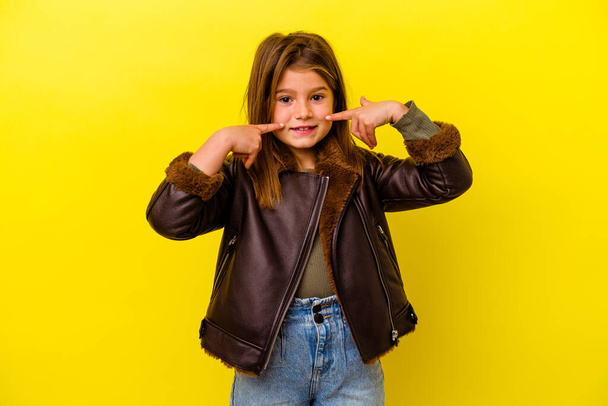 Маленькая кавказская девочка на желтом фоне улыбается, указывая пальцем на рот. - Фото, изображение