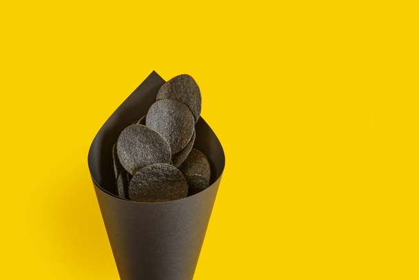 black potato chips in a black paper package on a yellow background, copy space, top view, chips made from natural ingredients, snack trend - Fotografie, Obrázek