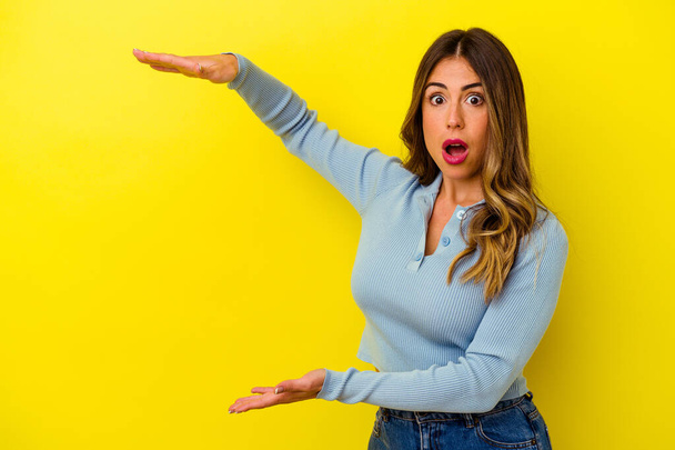Young caucasian woman isolated on yellow background shocked and amazed holding a copy space between hands. - Photo, Image
