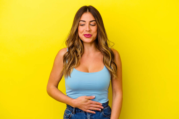 Young caucasian woman isolated on yellow background touches tummy, smiles gently, eating and satisfaction concept. - Foto, afbeelding