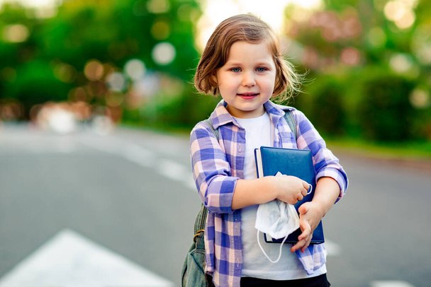 a schoolgirl is standing on the road in a plaid shirt and a medical mask, with a school denim backpack and a diary in her hands - Foto, Imagen