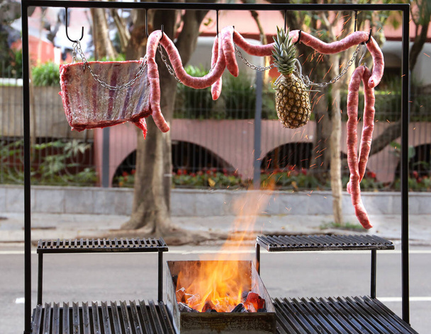 Argentine barbecue parrilla on the sidewalk in a street in Brazil. With fire and hanging sausages. - Photo, Image