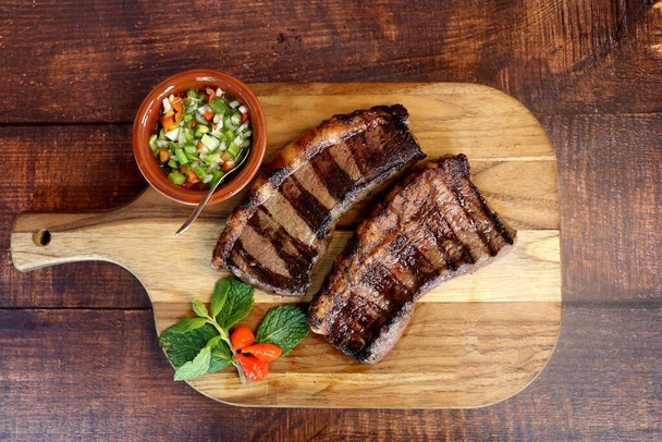 Picanha. Brazilian steak grilled in barbecue. Two slices of Picanha with green sauce, farofa, knife, vinagrete. Brazilian food. - Photo, Image
