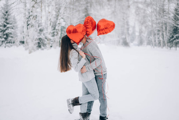 Joven pareja casada se besan en un parque nevado, escondiéndose detrás de globos en forma de corazón. - Foto, Imagen