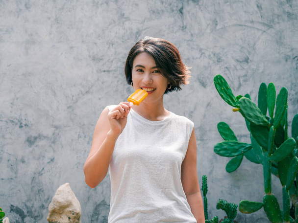 Smiling Asian woman short hair σε casual λευκό αμάνικο πουκάμισο με κίτρινη γρανίτα σε γκρι φόντο τσιμεντένιου τοίχου το καλοκαίρι. Γυναίκες που τρώνε γρανίτες, φρέσκια καλοκαιρινή εποχή. - Φωτογραφία, εικόνα