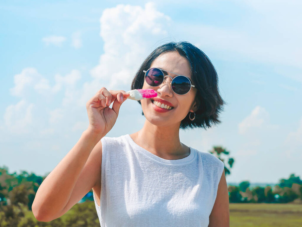 Smiling Asian woman short hair in casual white sleeveless shirt φορώντας γυαλιά ηλίου κρατώντας ροζ γρανίτα σε φόντο μπλε ουρανού το καλοκαίρι. Γυναίκες που τρώνε γρανίτες, φρέσκια καλοκαιρινή εποχή. - Φωτογραφία, εικόνα