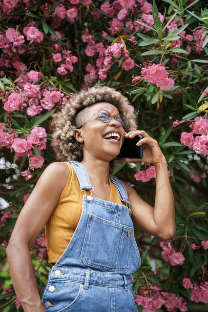 Mujer con estilo afroamericano sonriendo en una llamada telefónica rodeada de rosas rosadas en el jardín - Foto, Imagen