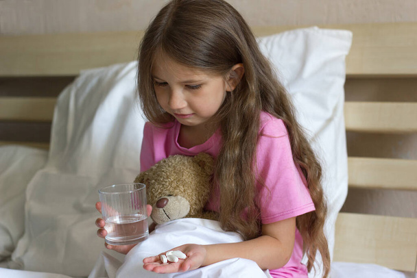 A cute sad sick little girl with blond hair in a pink T-shirt sits on the bed holding a glass of water, pills, medicines and a teddy bear  - Photo, Image