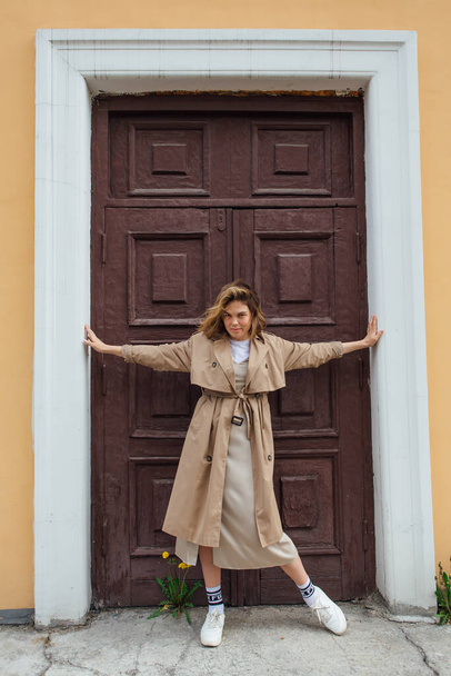 Young smiling millennial woman with wild hair dressed in an autumn coat posing near the door of an old building. - Foto, Imagen