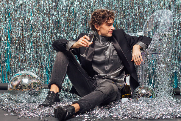 Tall attractive man sitting on the floor over the sparkle background and mirror disco ball holding glass of champagne. Party concept. - Foto, Imagen