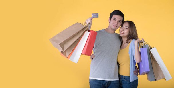 黄色の背景に隔離されたクレジットカードとショッピングバッグを持つアジアのカップル - 写真・画像