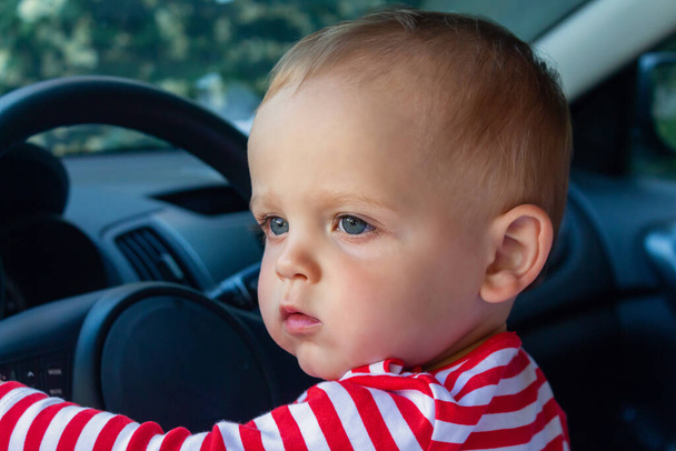Small caucasian baby boy as driver. Sitting in driver's seat in car. Travel with kids concept. Baby looking at something through the open car window.  - Fotó, kép