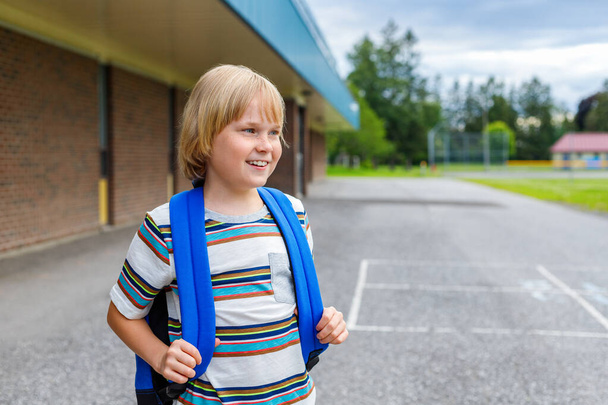 Kaunis hymyilevä lapsi seisoo koulun rakennuksen lähellä koulun pihalla. Pieni onnellinen blondi opiskelija kuljettaa sininen reppu. Takaisin kouluun ja koulutus käsite. - Valokuva, kuva