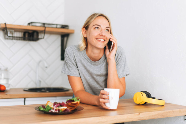 Молода усміхнена блондинка в жовтій футболці з чашкою чаю розмовляє на мобільному телефоні в руках на кухні вдома
 - Фото, зображення