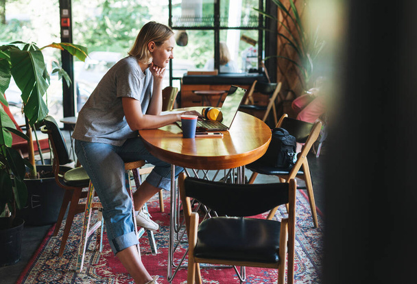 Mladá usměvavá blondýna žena na volné noze ve žlutých sluchátkách pracuje na notebooku na stole v kavárně - Fotografie, Obrázek