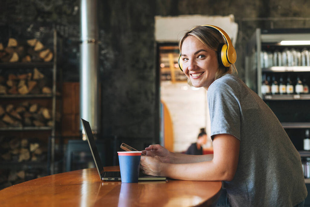 Mladá usměvavá blondýna žena na volné noze ve žlutých sluchátkách pracuje na notebooku na stole v kavárně - Fotografie, Obrázek