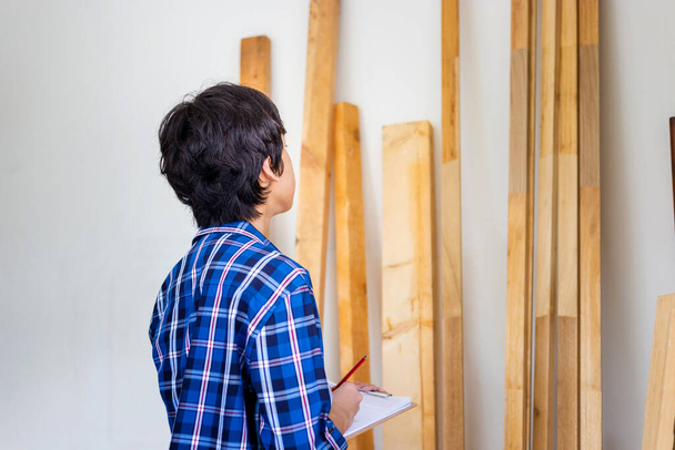 Mladý tesař kontroluje skladiště dřeva v továrně. Muž počítání dřeva trámy zásob dřeva v truhlářské dílně - Fotografie, Obrázek