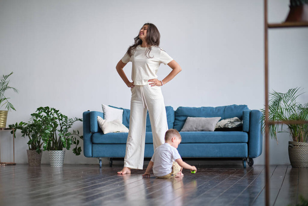 Onnellinen tietoinen yksinhuoltajaäiti tekee aamuharjoituksia joogassa aiheuttaa kanssa lapsi pelaa kotona. Aamuharjoituksia pyjamassa. lapsi häiritsee harjoittelua - Valokuva, kuva