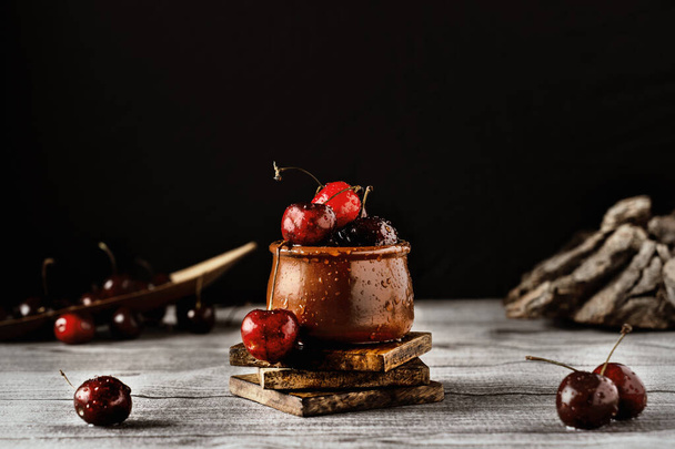 A beautiful wallpaper with a clay pot full of cherries that has water dripping down it on a wooden table - Фото, изображение