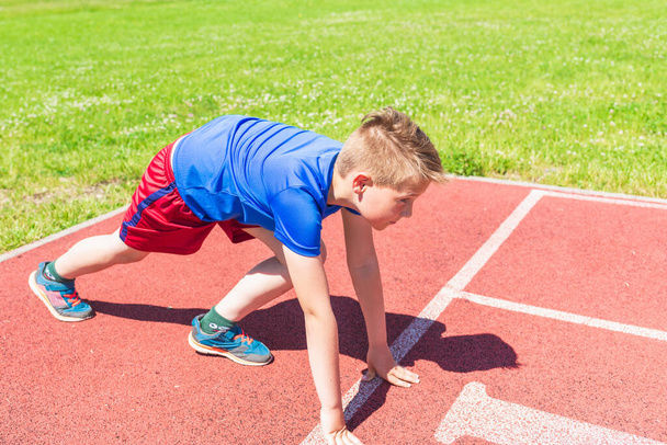 Caucasian schoolboy prepare to start running on a red track.young boy in starting position ready for running. Healthy lifestyle.Summer day. - Photo, Image
