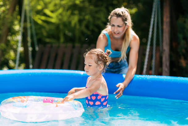 Nuori äiti opettaa pienen tyttärensä uimaan puutarhan uima-altaassa puhallettavan ympyrän avulla. - Valokuva, kuva
