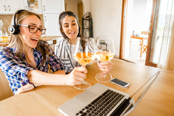 İki mutlu genç kadın masada oturmuş dizüstü bilgisayarlarıyla şampanya ya da beyaz şarap kadehleriyle kadeh kaldırıyor. Sosyal uzaklık ve video konferans teknolojisi sayesinde yeni normal kutlama çevrimiçi - Fotoğraf, Görsel