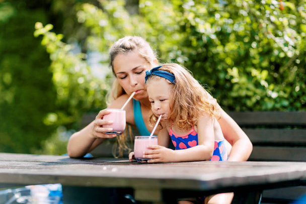 Una bella ragazza e la sua bambina bevono un frullato rinfrescante mentre sono seduti su una panchina in giardino - Foto, immagini
