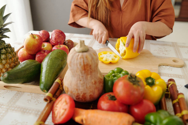 Close-up image of young woman sitting at kitchen table and cutting yellow bell pepper - Foto, Imagem