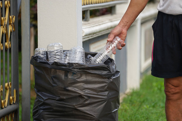 Рука кидає пластикову пляшку в мішок для сміття перед парканом будинку (вибірковий фокус). Концепція сортування та переробки побутових відходів
                                                              - Фото, зображення