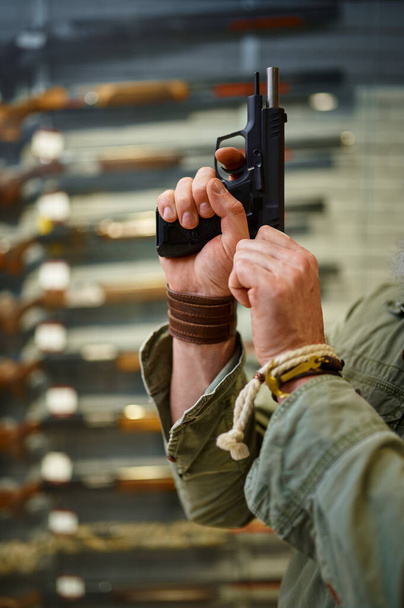 Bearded man twitches the pistol bolt in gun store. Weapon shop interior, ammo and ammunition assortment, firearms choice, shooting hobby and lifestyle - Photo, Image