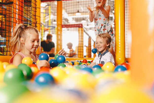 Счастливые дети играют в бассейне. Мальчики и девочки играют с разноцветными пластиковыми шариками в большом сухом бассейне для гребли в игровом центре. Школьники развлекаются на детской площадке в крытом парке  - Фото, изображение