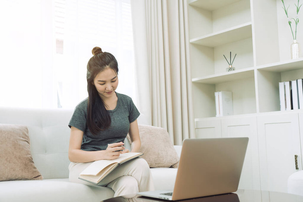 Mujer asiática se sienta en el sofá mientras toma notas de la pantalla de su computadora mientras trabaja o estudia en línea en casa. - Foto, imagen