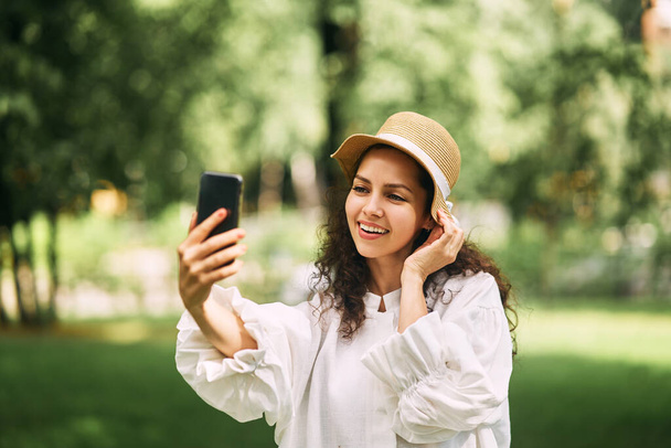 Jong mooi meisje met een hoed maakt een selfie op haar telefoon in het park. Hoge kwaliteit foto - Foto, afbeelding
