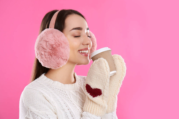 Beautiful young woman in earmuffs with cup of drink on pink background - Photo, image