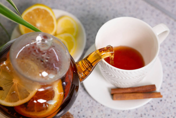 Una bebida de té caliente con limón y naranja, una tetera y una taza blanca, servidas con delicioso limón y canela. Té fragante y caliente. Etapa 2 - Foto, imagen