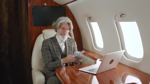 Feliz anciano rico cuenta el dinero mientras vuela en un jet privado. Exitoso sonriente millonario masculino se regocija en el beneficio, cuenta dólares en efectivo, durante un vuelo de primera clase - Imágenes, Vídeo
