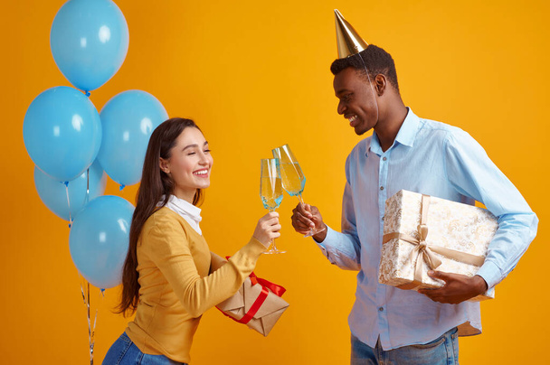 Couple drôle dans des casquettes tenant des verres de boissons et des boîtes-cadeaux, fond jaune. Jolie femme a obtenu une surprise, événement ou fête d'anniversaire, décoration de ballons - Photo, image