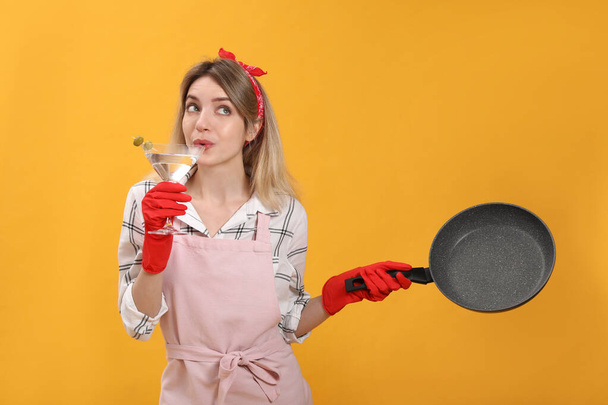Young housewife with frying pan and glass of martini on yellow background - Photo, image