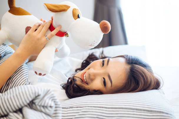 Young happy and positive Asian woman smiling in casual clothing while holding a dog doll in cozy living room on bed against white background - Foto, Bild